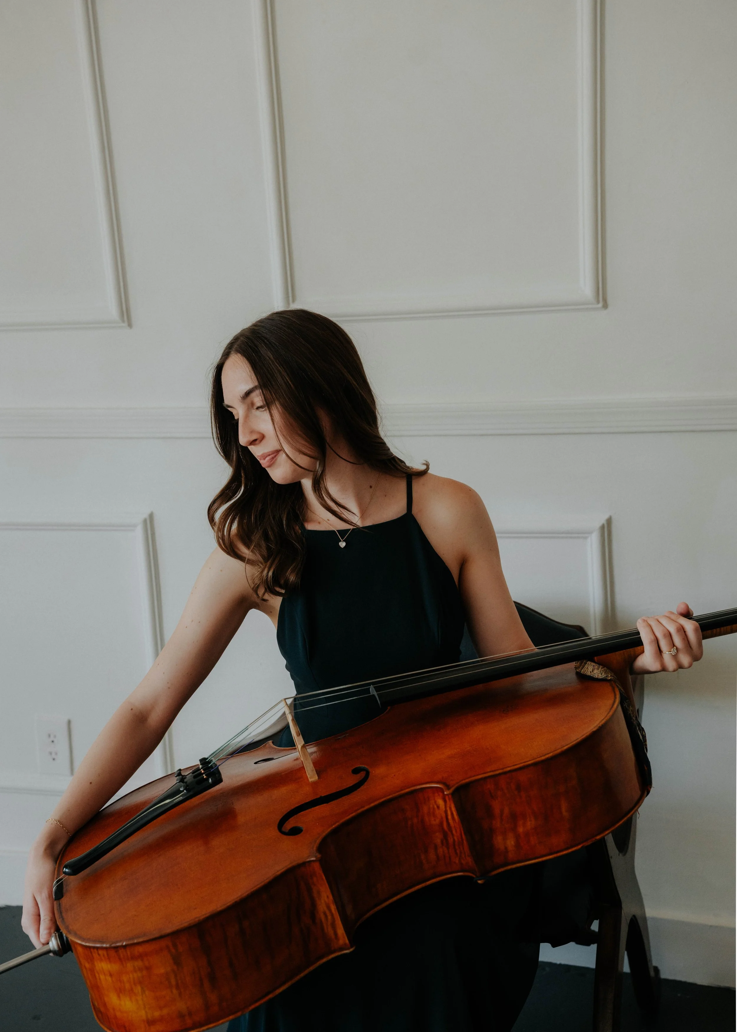 A young woman with long brown hair wearing a black dress playing a large cello in a room with white paneled walls.