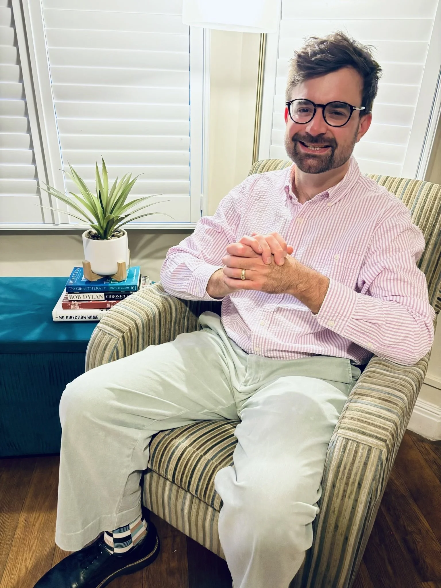 A man with glasses, a beard, and short brown hair, smiling and seated in a green striped armchair. He is wearing a pink and white striped shirt, white pants, and black shoes. There is a small table with a stack of books and a potted plant behind him, and windows with white shutters in the background.