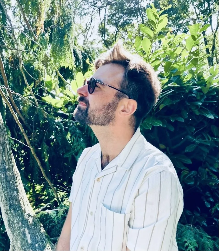 A man with a beard and sunglasses standing outdoors in a green, leafy environment, wearing a white striped shirt and looking to the side.