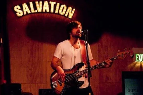 A man playing a bass guitar and singing into a microphone on stage with a wood-paneled wall behind him and a lit-up 'SALVATION' sign above.
