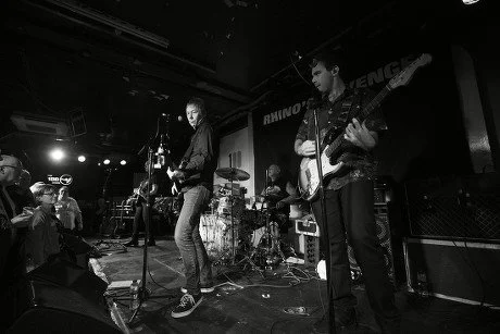 A band performs live on stage in a dimly lit venue with instruments, microphones, and a drummer in the background.