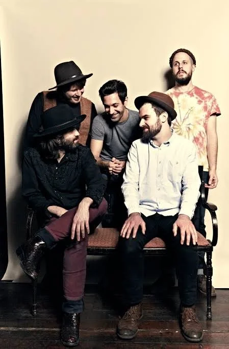 Five men gathered around a vintage wooden bench against a plain beige wall, engaging in conversation and laughter.