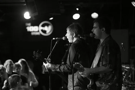 Two musicians performing on stage with a microphone, in a venue with an audience and stage lighting.