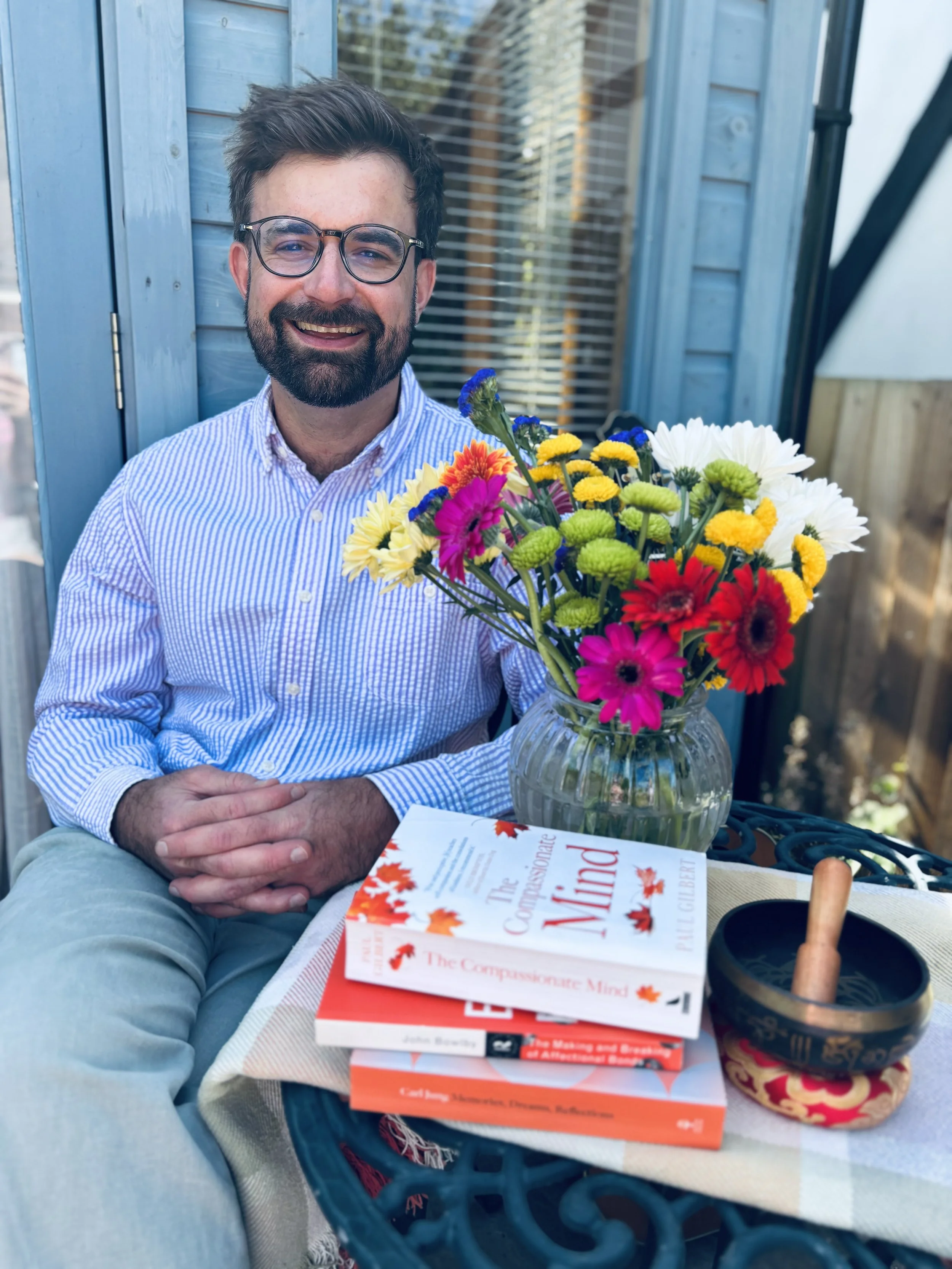 A man with glasses and a beard sitting outdoors, smiling, holding a bouquet of colorful flowers. In front of him are three stacked books about compassion and empathy, and a cup with a spoon inside.