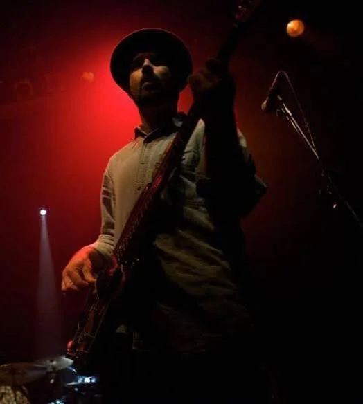 A musician wearing a hat and a light-colored shirt playing an electric guitar on stage, illuminated by red lighting with a dark background.