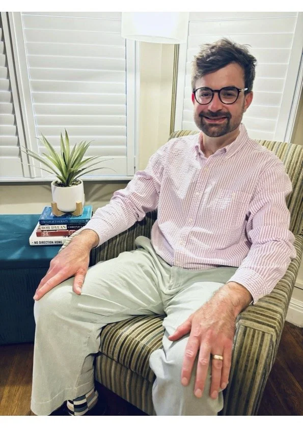 A man with glasses and a beard sitting on a striped armchair, smiling, wearing a light pink striped shirt and light-colored pants, with a gold wedding ring on his left hand. Behind him are white window shutters, and a small table with a potted plant and a stack of books.