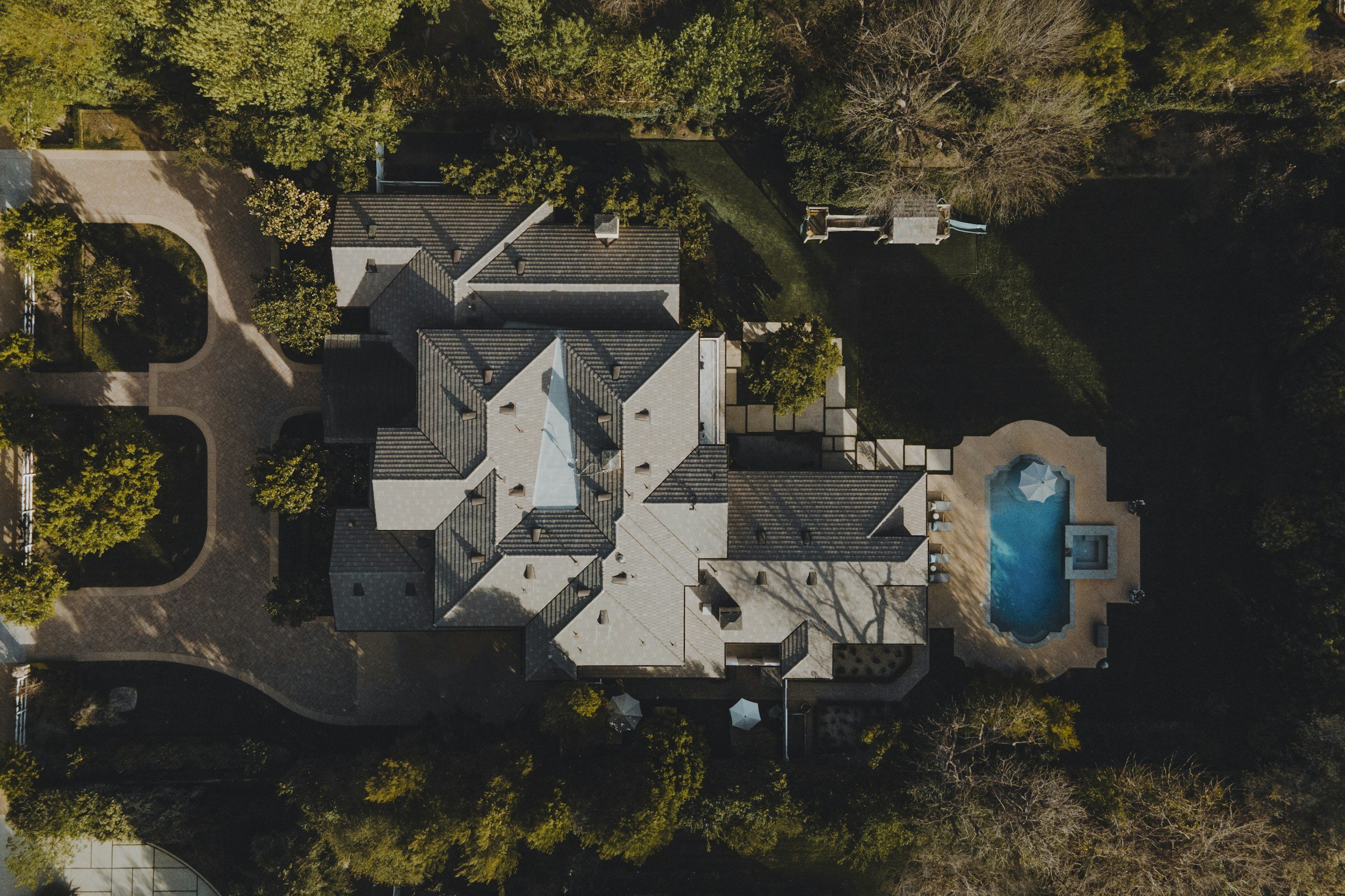 An aerial view of a large residential house with a gray roof, surrounded by trees, with a backyard that includes a swimming pool, a patio, and a playground.