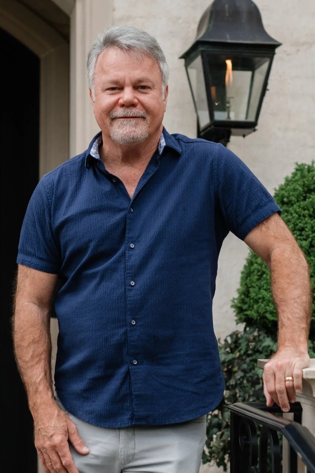 A middle-aged man with gray hair and a beard, wearing a navy blue short-sleeve button-up shirt and light-colored pants, standing outdoors near a house with a lantern-style outdoor light fixture on the wall and greenery in the background.