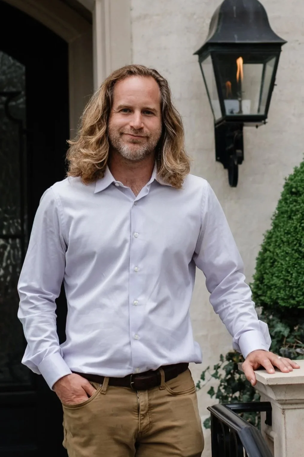 A man with long, wavy hair and a beard, wearing a white button-down shirt and beige pants, standing outdoors beside a black lantern-style wall light and a white stone railing with greenery.