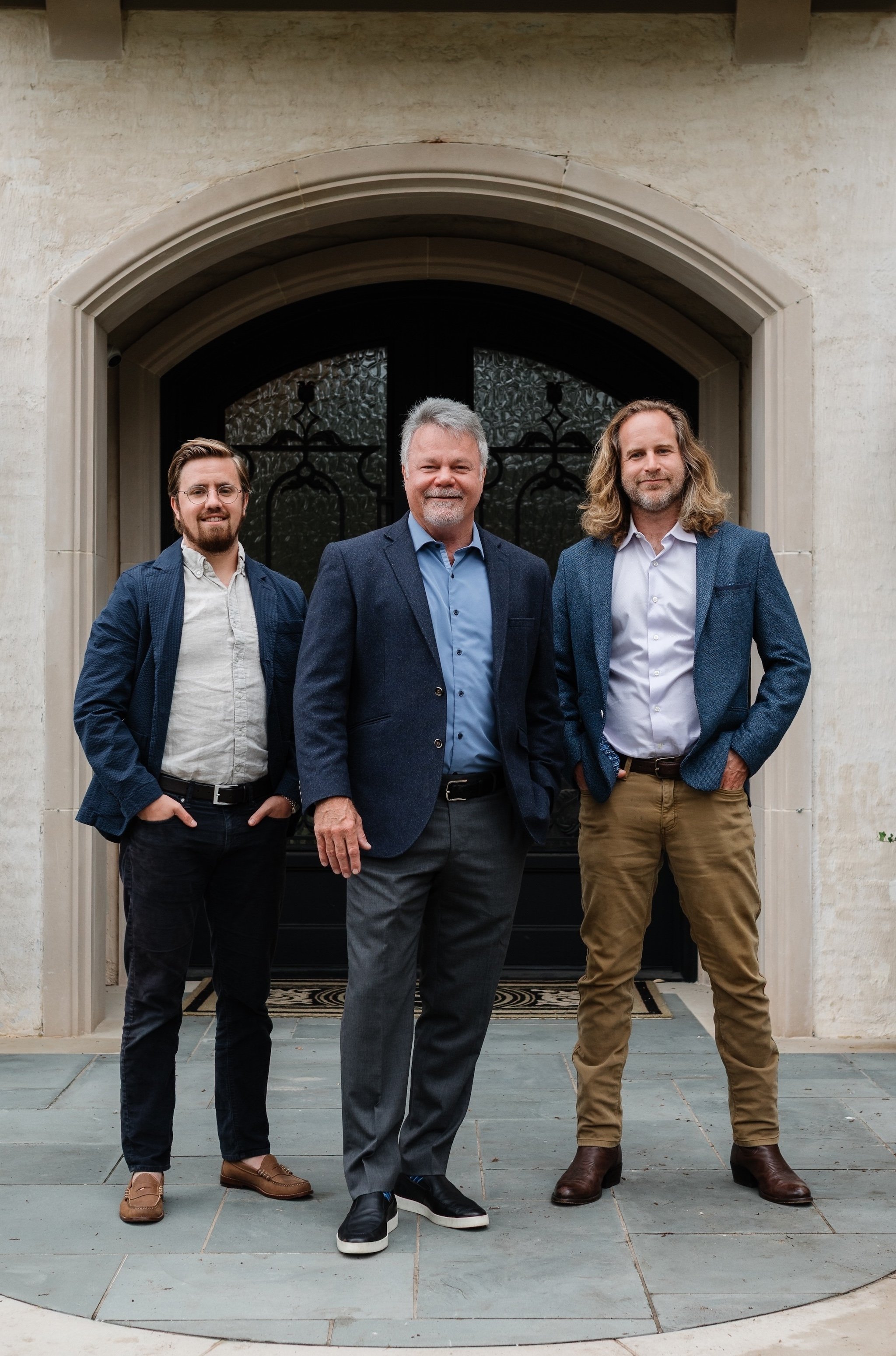 Three men in business casual attire standing in front of a decorative black iron gate and a beige stone building. The man in the center has gray hair and a beard, wearing a blue blazer and gray pants. The man on the left has glasses, a beard, and is wearing a navy jacket, light-colored shirt, and dark pants. The man on the right has long blond hair, a beard, and is wearing a blue blazer, white shirt, and tan pants.
