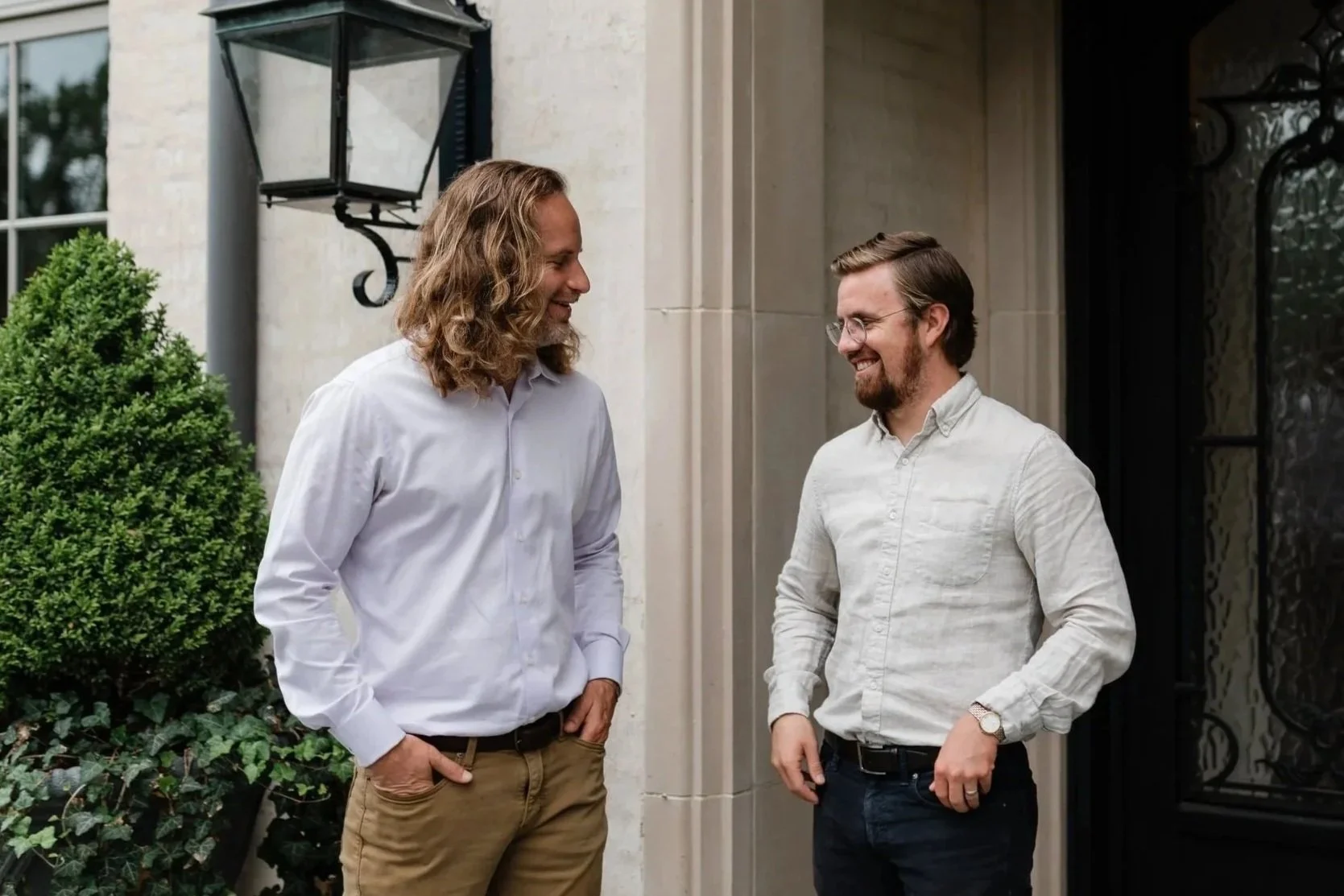 Two men standing outside, smiling and engaging in conversation, near a doorway with a large black metal door and a lantern-style light fixture mounted on the wall. One man has long, wavy hair and a beard, wearing a white shirt and tan pants. The other has short hair, glasses, a beard, and is wearing a light-colored shirt and dark pants.