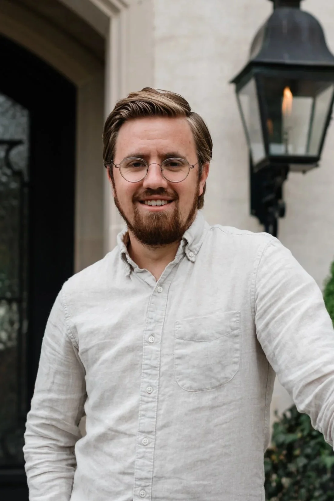 A young man with glasses and a beard wearing a light-colored button-up shirt standing outside near a house with a black outdoor lantern in the background.