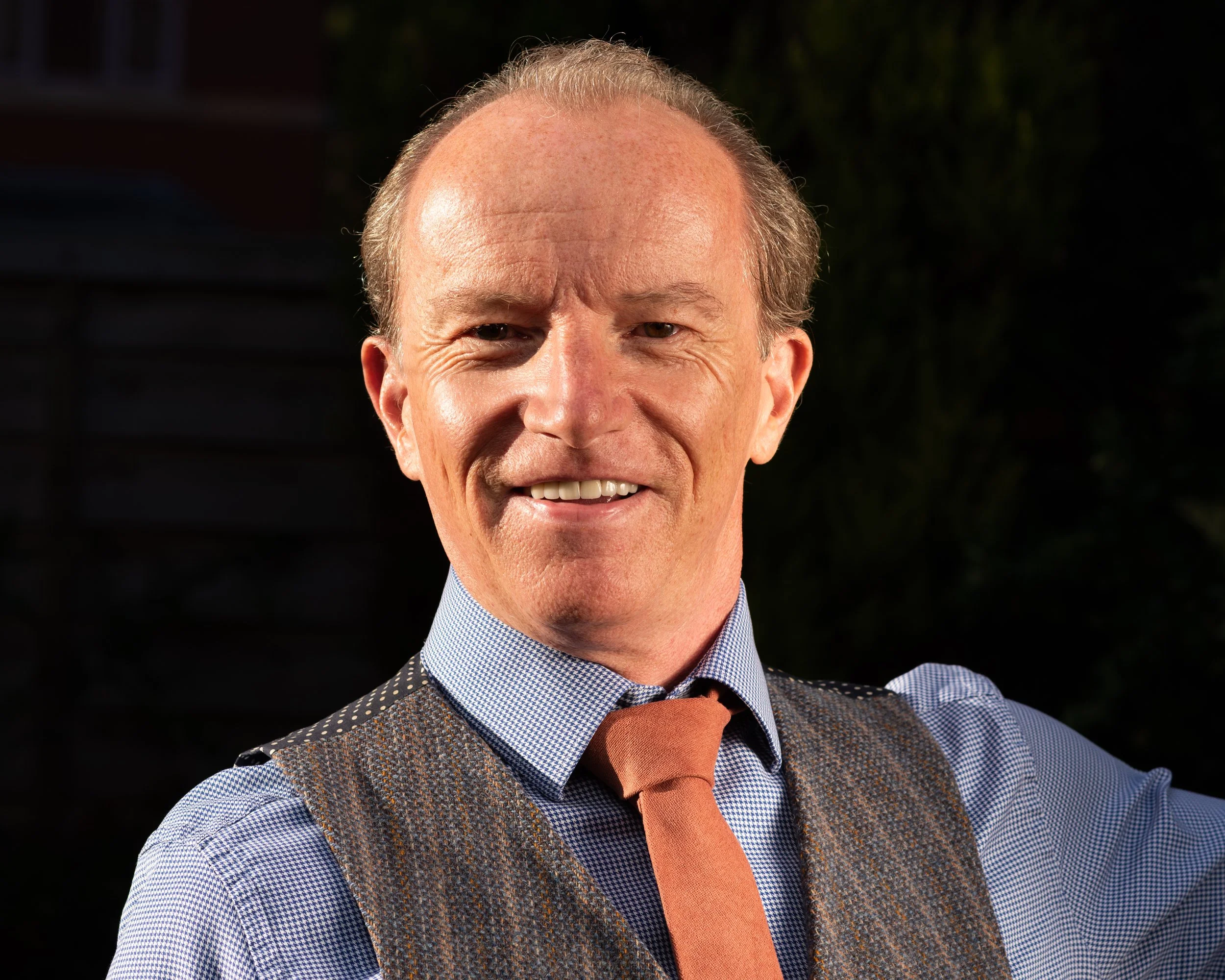 Close-up of a smiling middle-aged man wearing a blue checkered shirt, orange tie, and brown vest, outdoors during evening or night.
