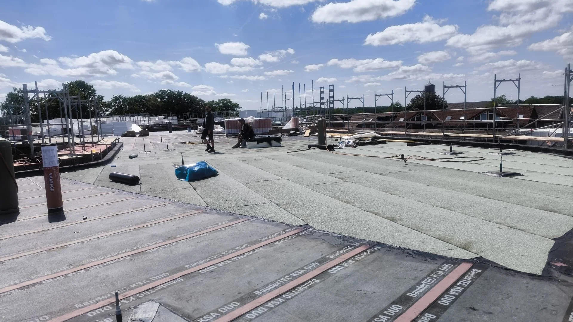 Auf einem flach gedeckten Dach befinden sich Baumaterialien, Gerüstteile, Werkzeuge und drei Männer, die bei Arbeiten auf dem Dach im Sonnenschein sind. Im Hintergrund sind Häuser mit roten Dächern und ein blauer Himmel mit Wolken sichtbar.