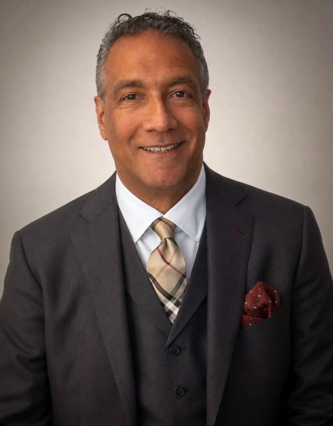 A smiling man wearing a dark gray suit, white shirt, plaid tie, and a red pocket square, standing against a neutral background.