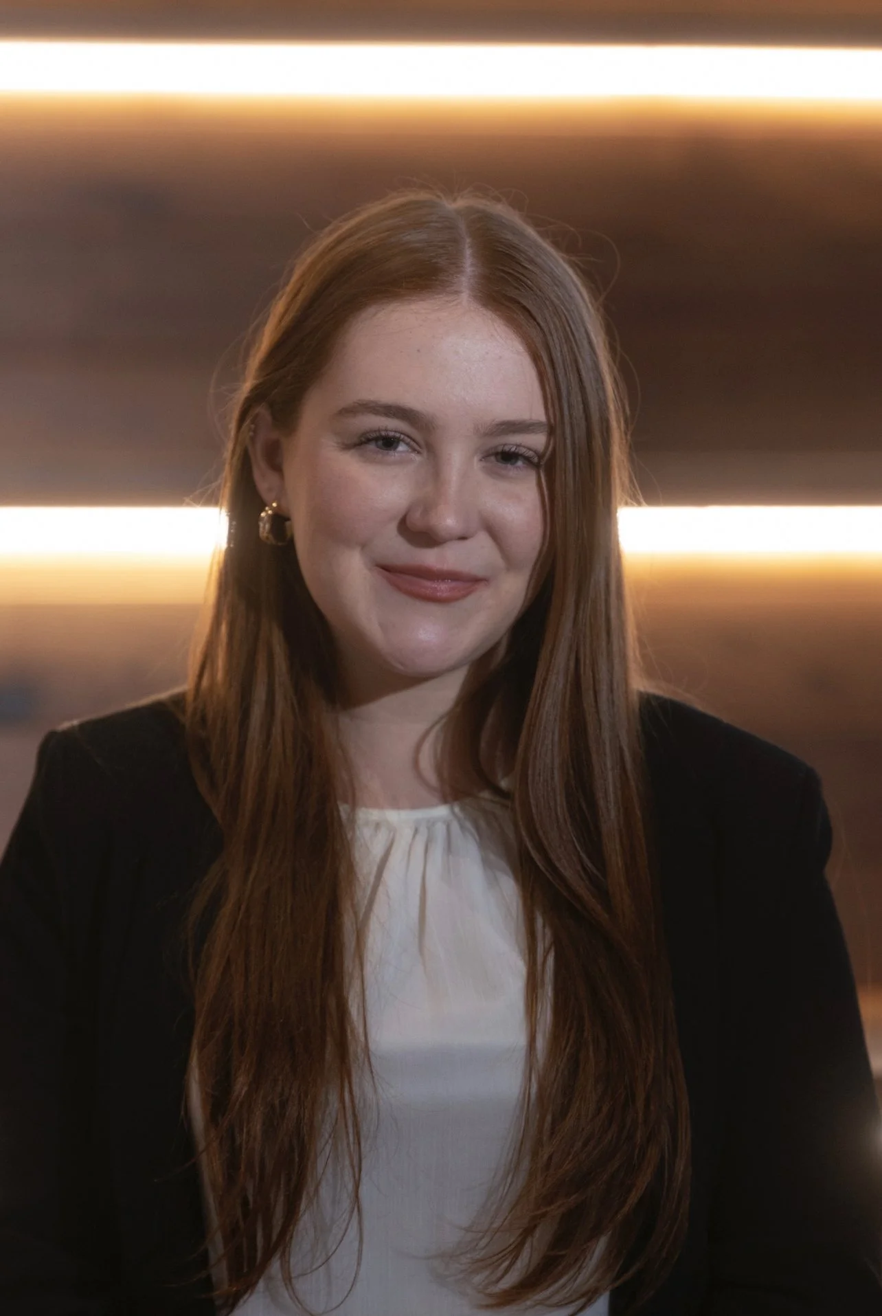 A young woman with long red hair, wearing gold hoop earrings, a black blazer, and a white blouse, smiling in front of a background with horizontal bright light lines.