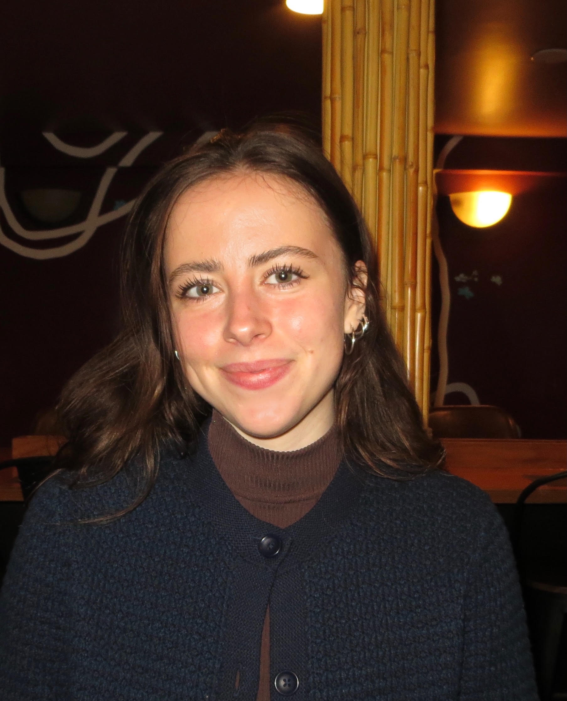 Close-up of a woman with long brown hair, green eyes, and earrings smiling in a dimly lit indoor setting with a wicker divider and wall lights in the background.