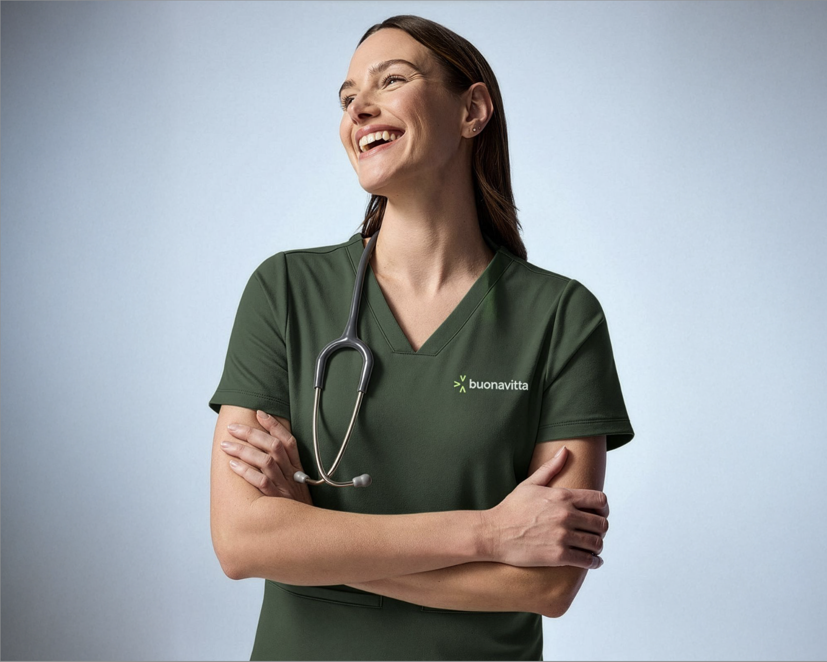 Mujer enfermera de pie con brazos cruzados, usando uniforme verde con logo de 'buonavita' y un estetoscopio alrededor del cuello, sonriente y mirando hacia la derecha, en un fondo de color gris claro.