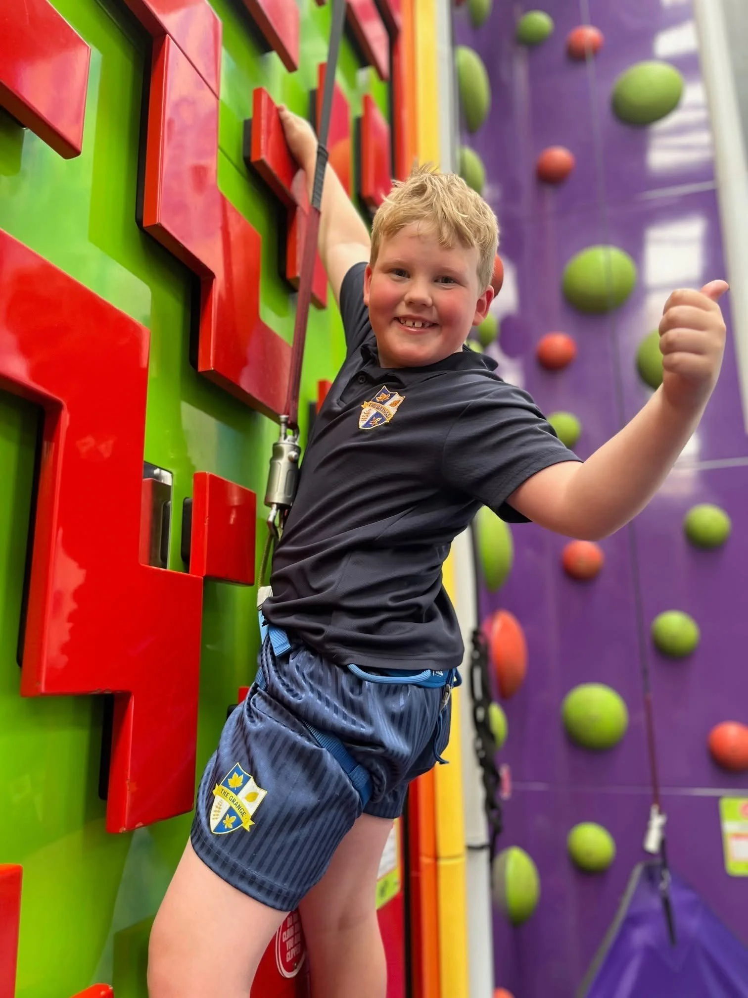 Smiling boy with thumbs up on a colourful climbing wall with rope harness