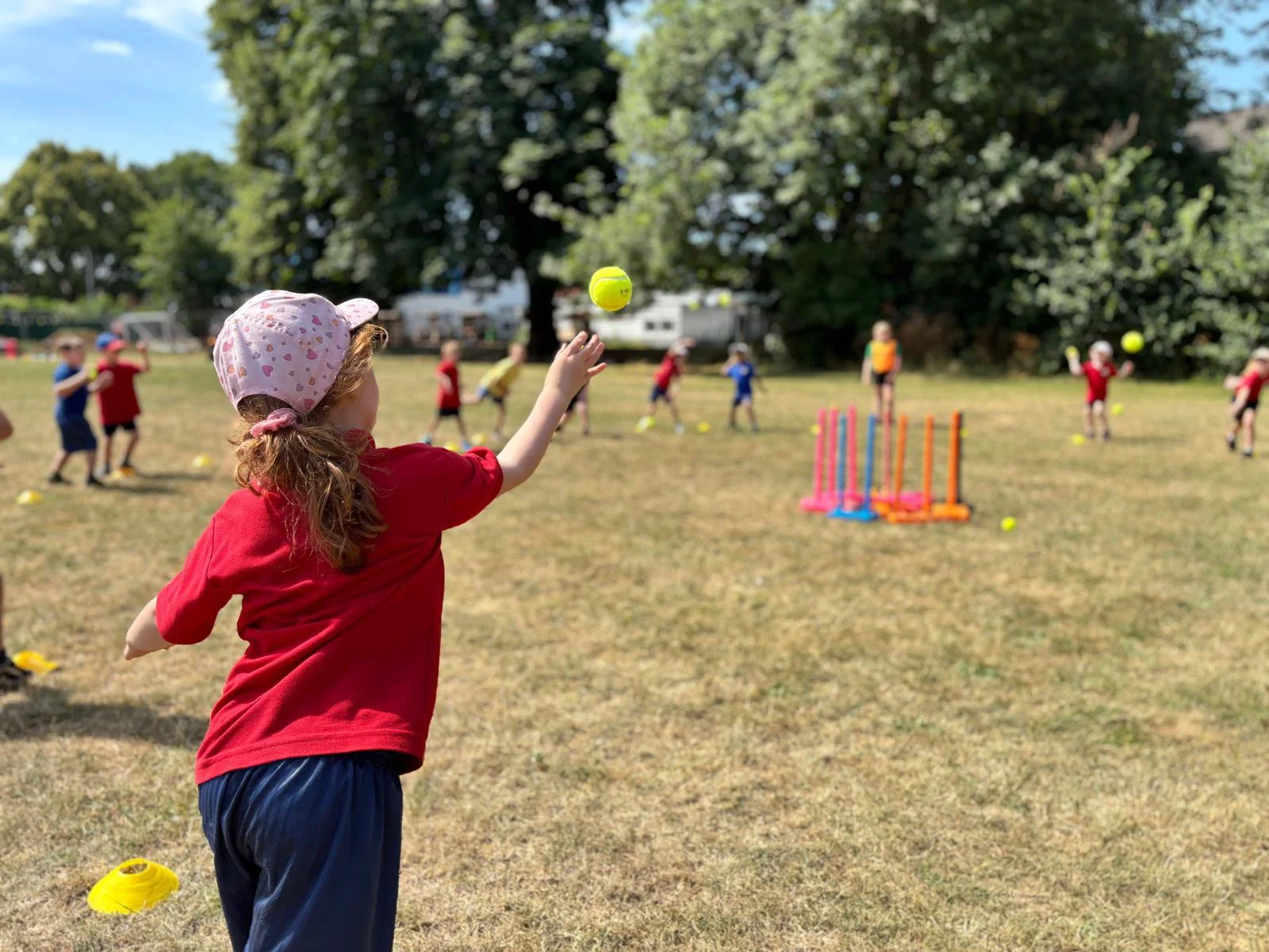 A young girl in a pink hat with love hearts throwing a yellow tennis ball in a primary school cricket game