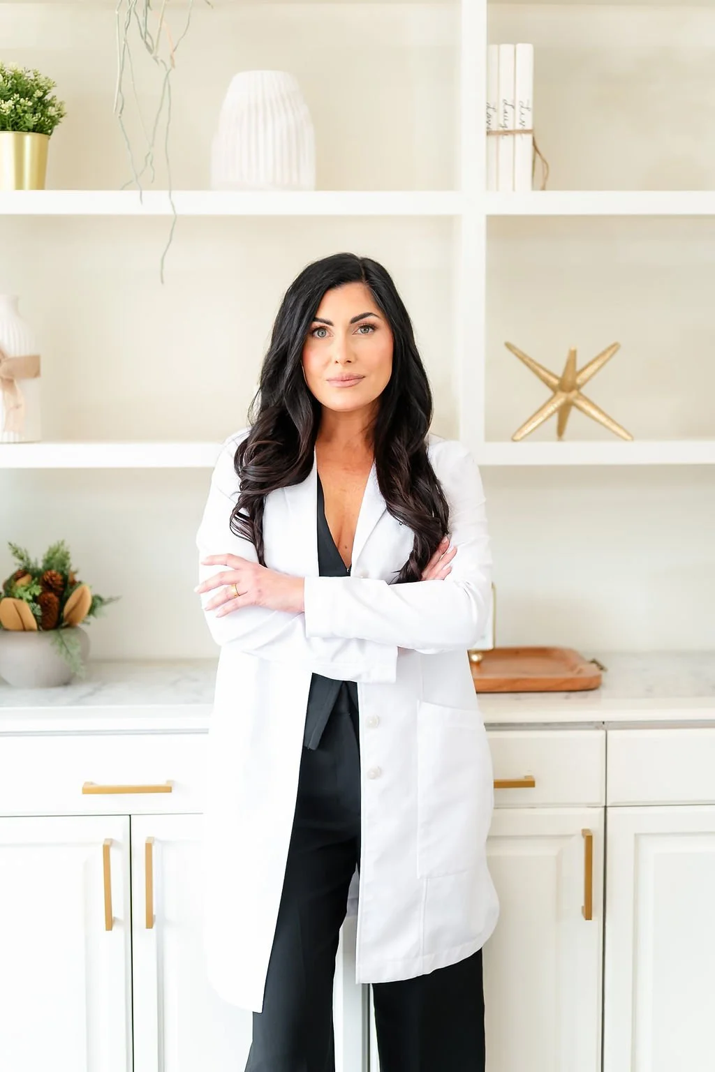 A woman with long black hair, wearing a white lab coat over black clothes, standing with arms crossed in a modern, light-colored kitchen with decorative items on the shelves.