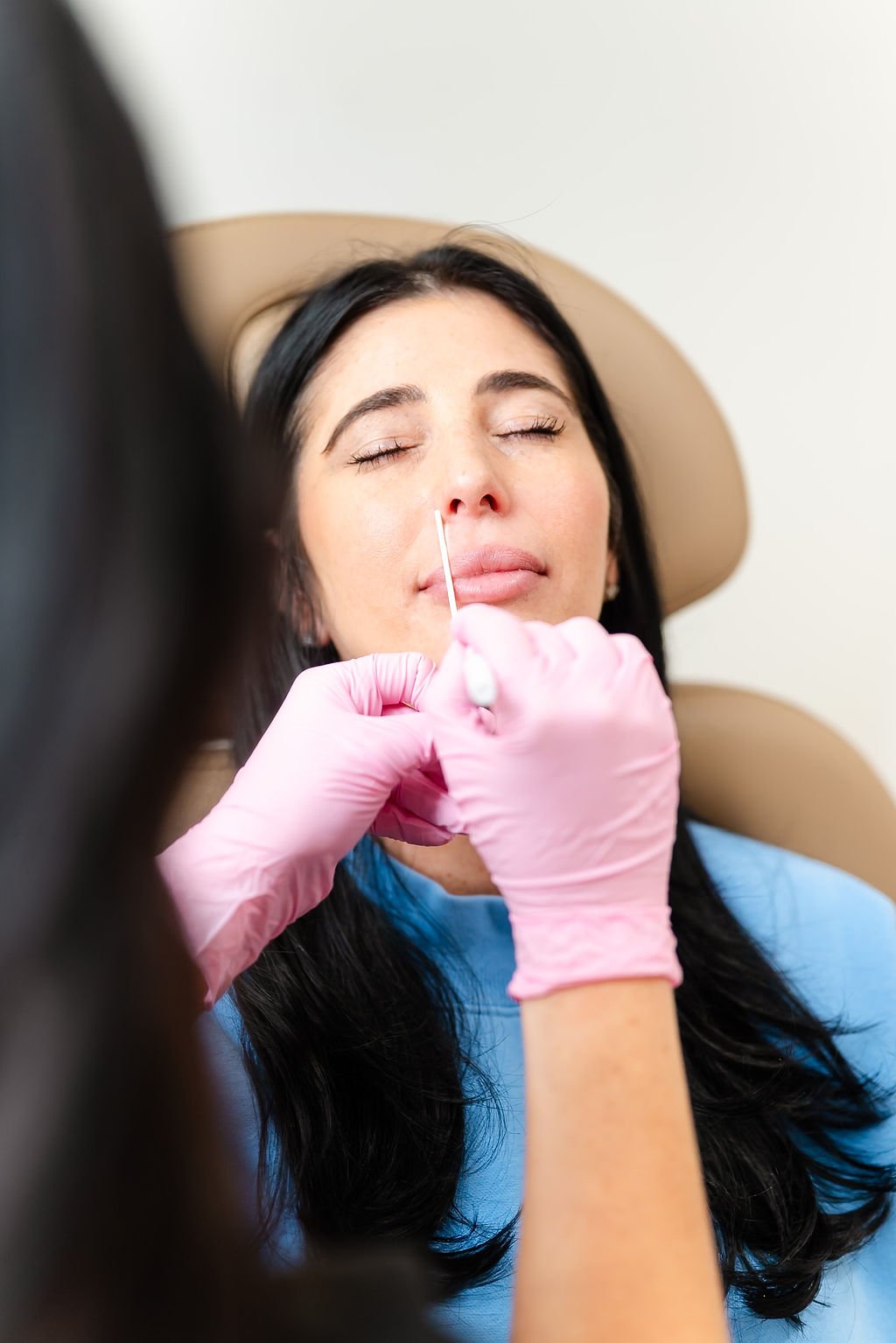 A woman receiving a cosmetic procedure, possibly a facial or skin treatment, in a medical setting with a healthcare professional using a cotton swab.