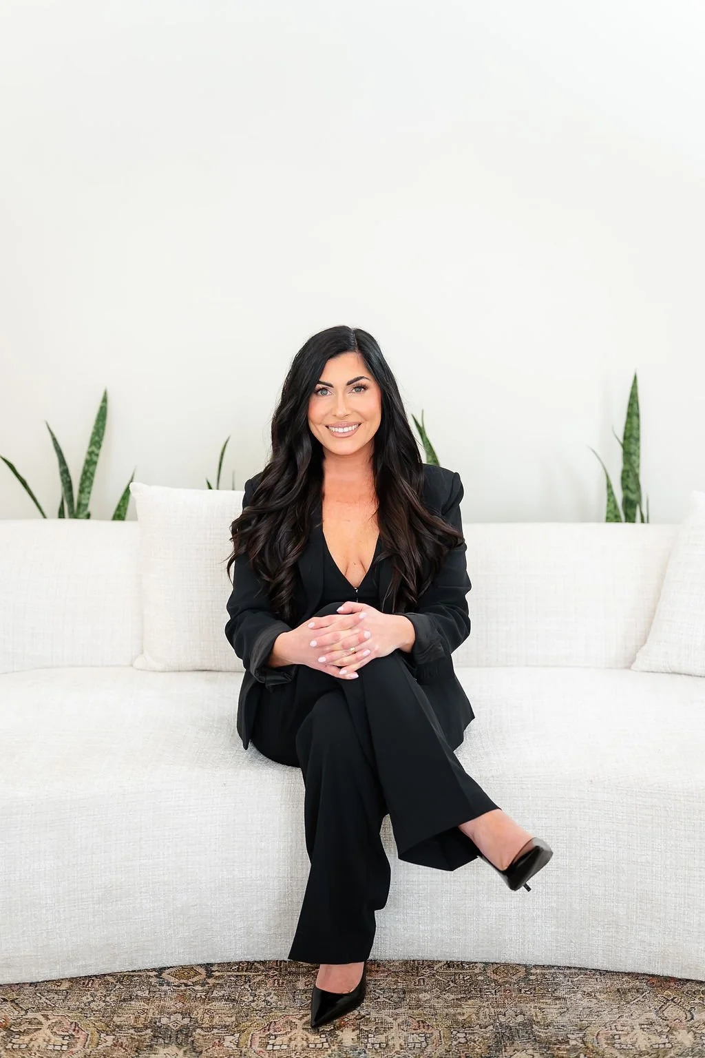 A woman with long dark hair in a black suit smiling and sitting on a white sofa with green plants in the background.