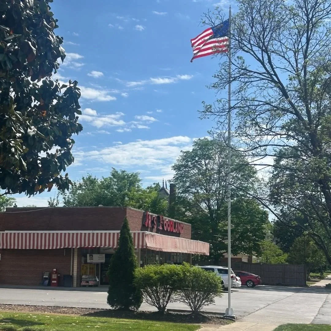 A storefront with a red and white striped awning, a flagpole with the American flag, surrounded by trees and parked cars, on a sunny day with blue sky and some clouds.
