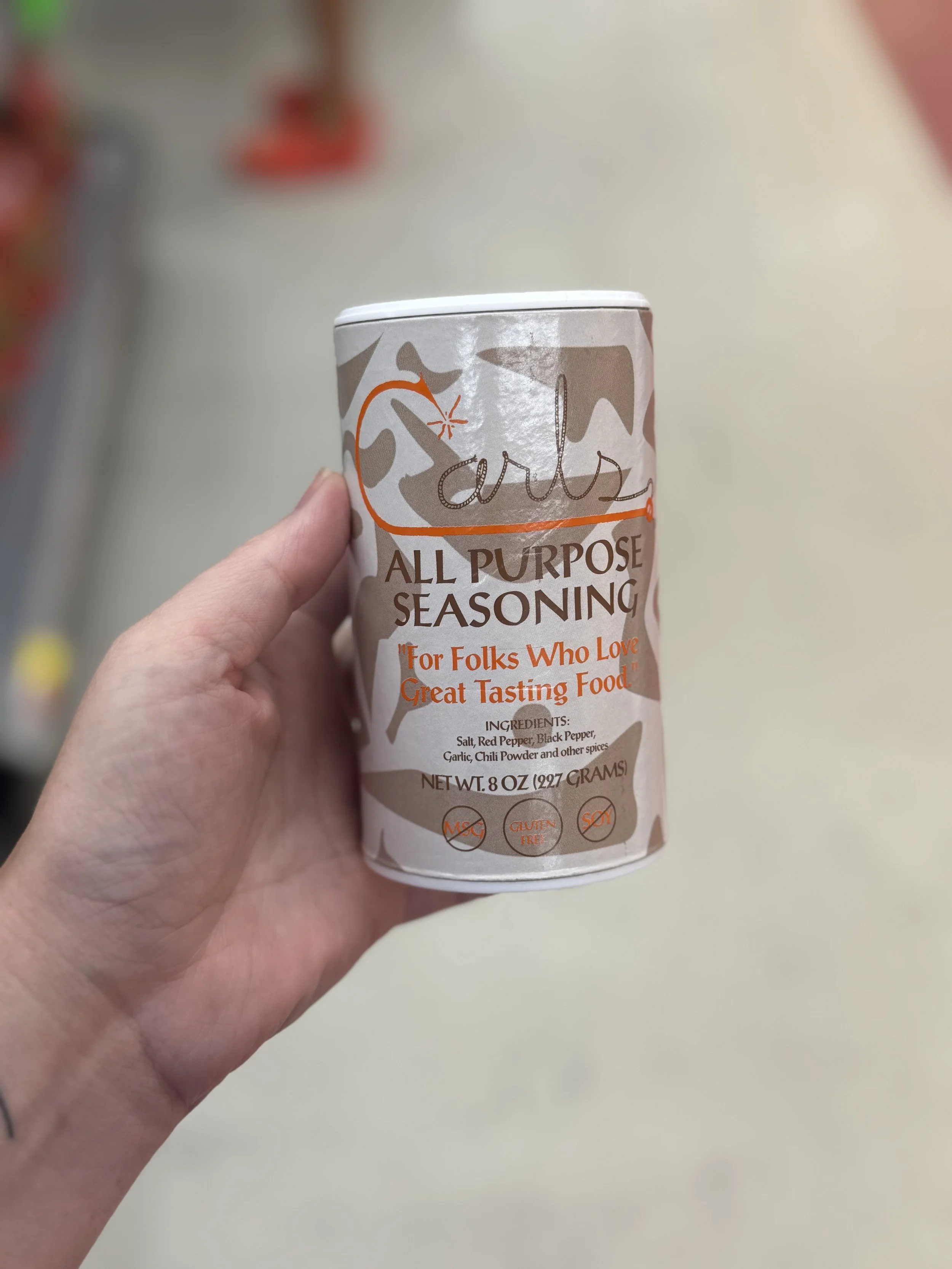 A hand holding a can of all purpose seasoning with a beige and orange label that has the word 'carl's' and 'ALL PURPOSE SEASONING' printed on it, along with a tagline 'For Folks Who Love Great Tasting Food' and ingredients list, against a blurred background.