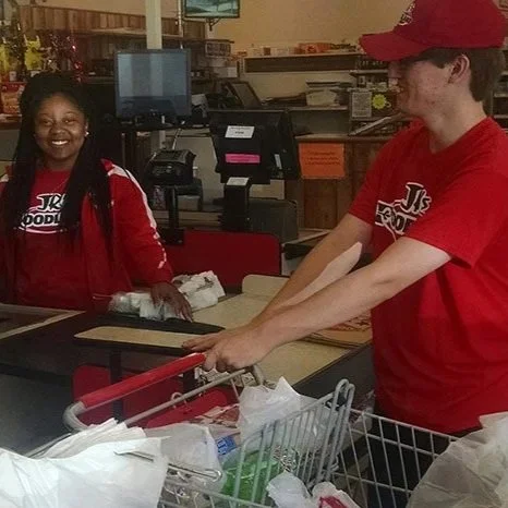 A young man and young woman shopping at a grocery checkout counter, both wearing red shirts with a logo, with the man pushing the cart and the woman smiling.