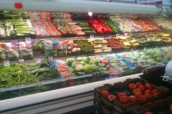 Refrigerated grocery store produce section with tomatoes, cucumbers, and various vegetables.