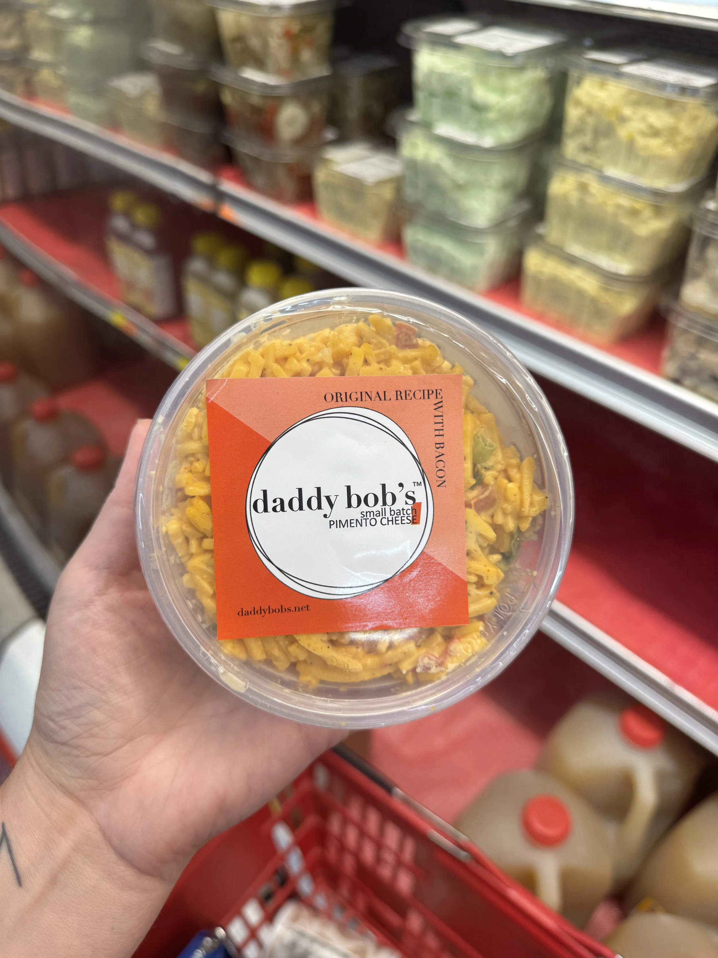 A hand holding a plastic container of macaroni and cheese with bacon. The container has a label from Daddy Bob's, featuring a black and white circle logo and the text "Original Recipe with Bacon". Shelves with various food items can be seen in the background at a grocery store.