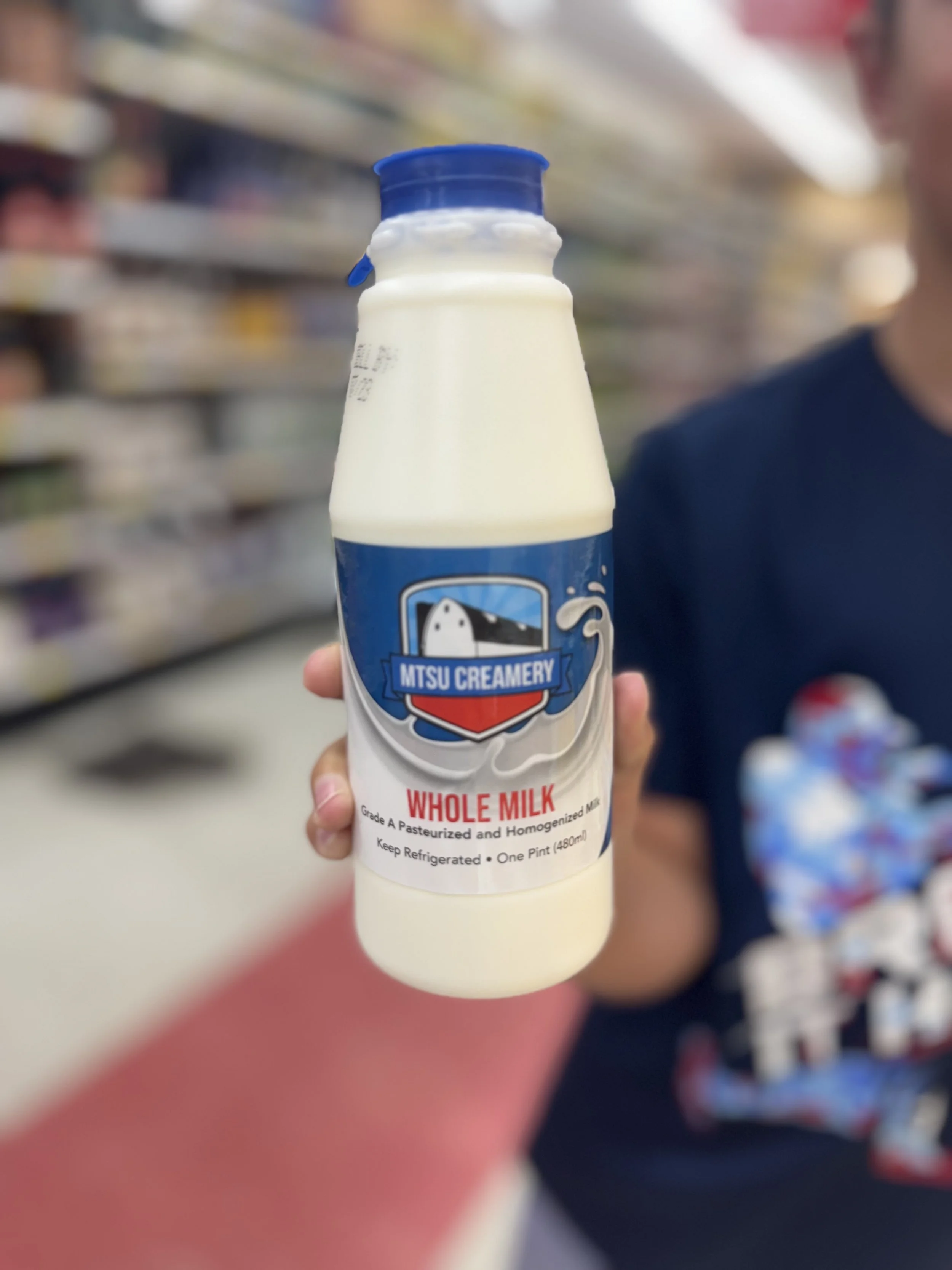 A person in a dark shirt holding a bottle of whole milk in a grocery store aisle.
