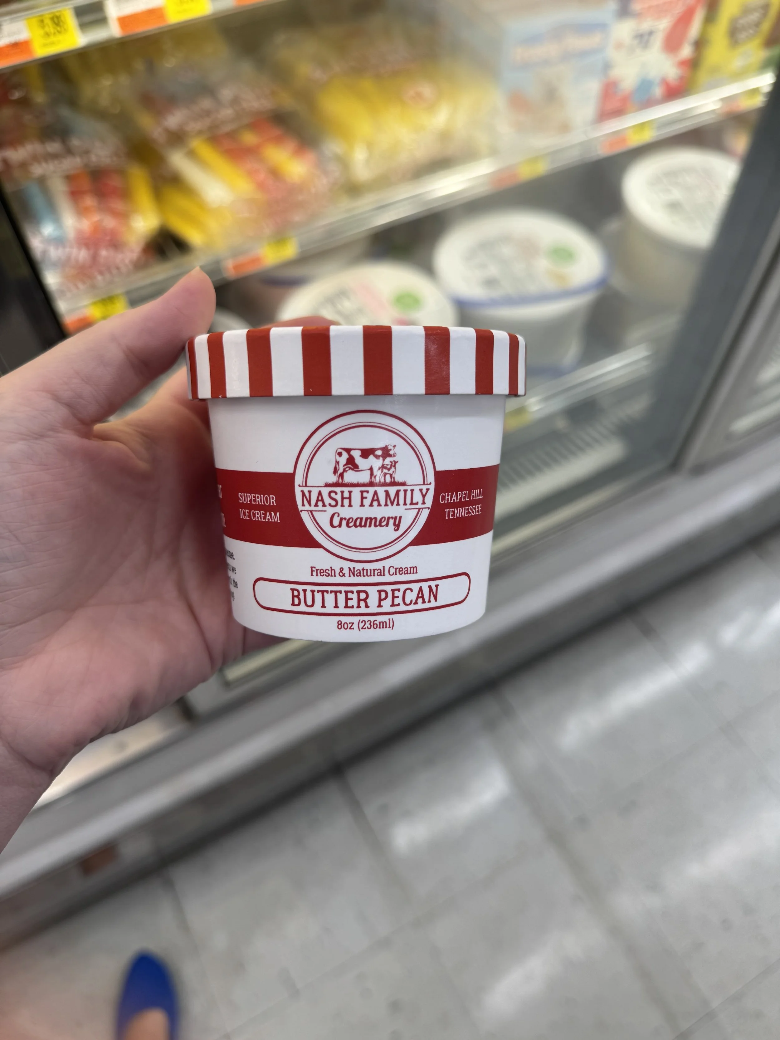 Person holding a cup of Nash Family Creamery Butter Pecan ice cream in front of a grocery store freezer with various ice cream flavors.