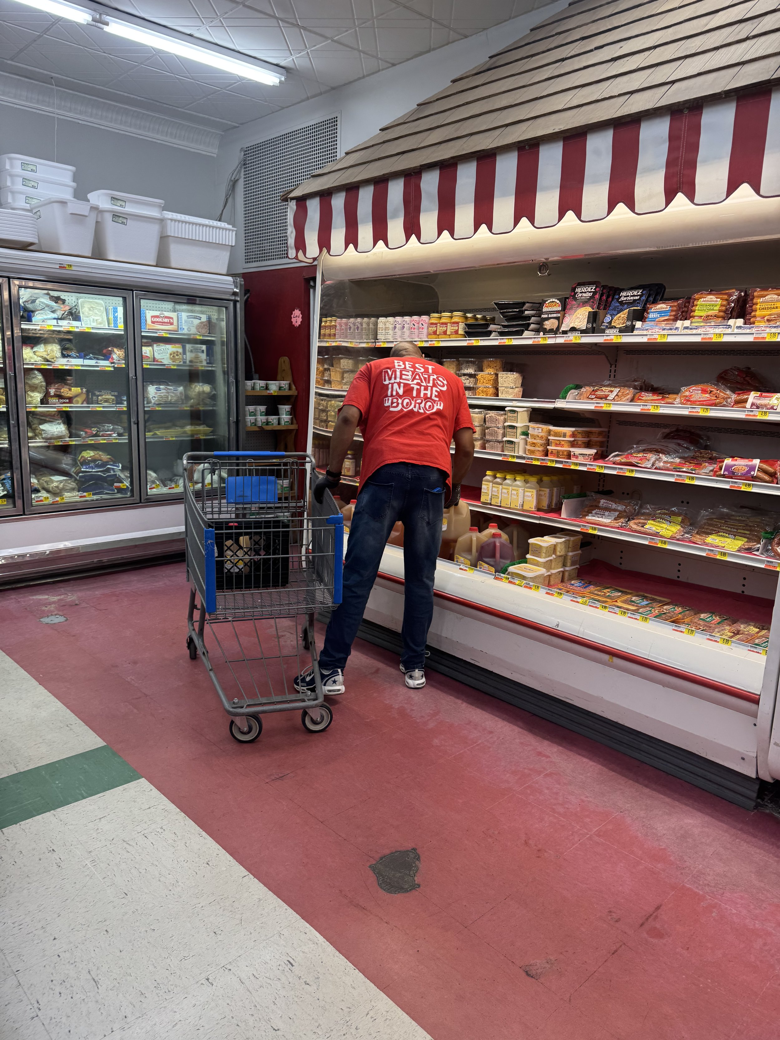 A man pushing a shopping cart in a grocery store aisle, reaching for items on a shelf filled with packaged foods and meats. The store has red, white, and green colored tile flooring and a refrigerated section on the left.