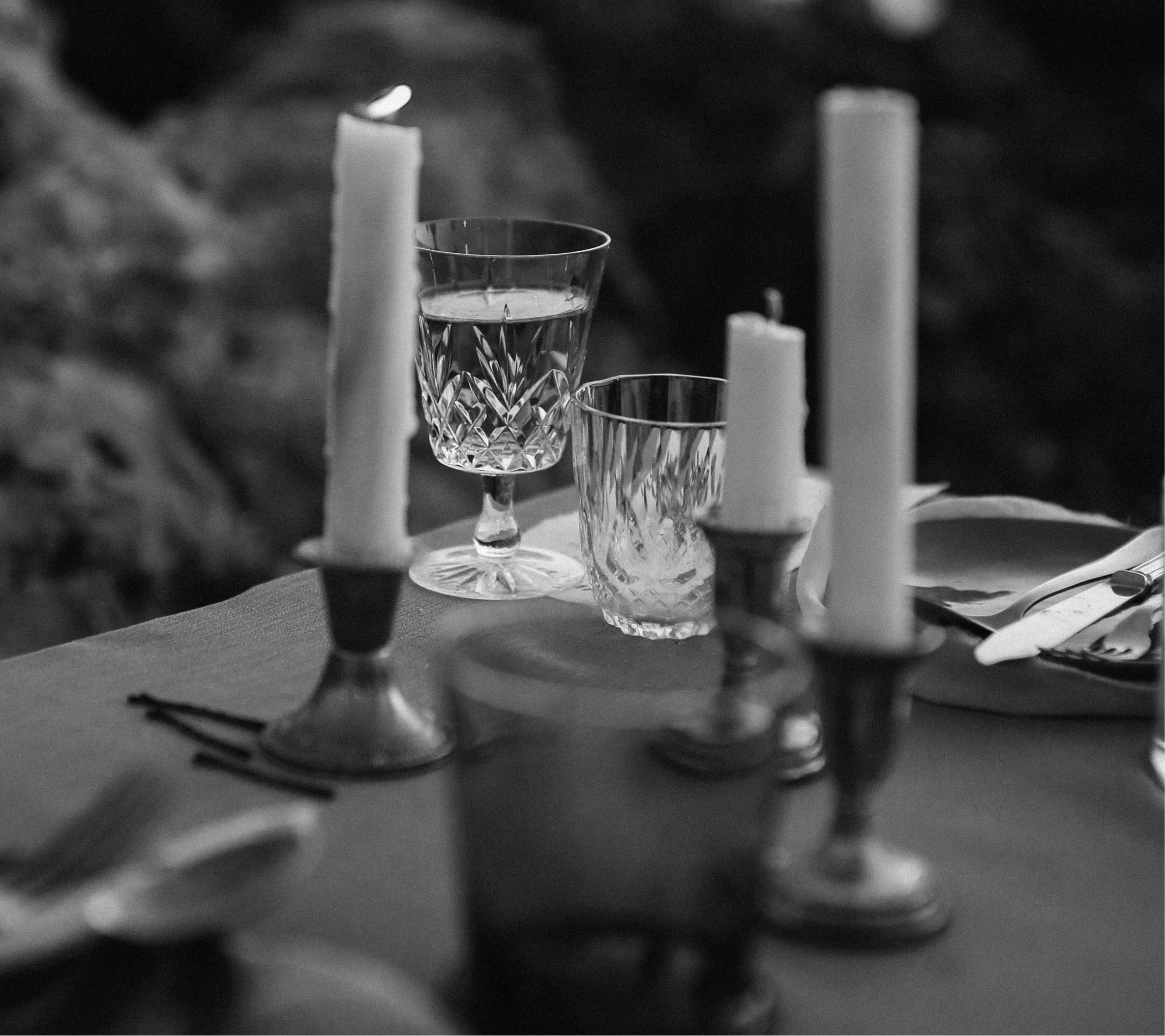 Black and white photo of crystal glasses with a drink, surrounded by candles and tableware.