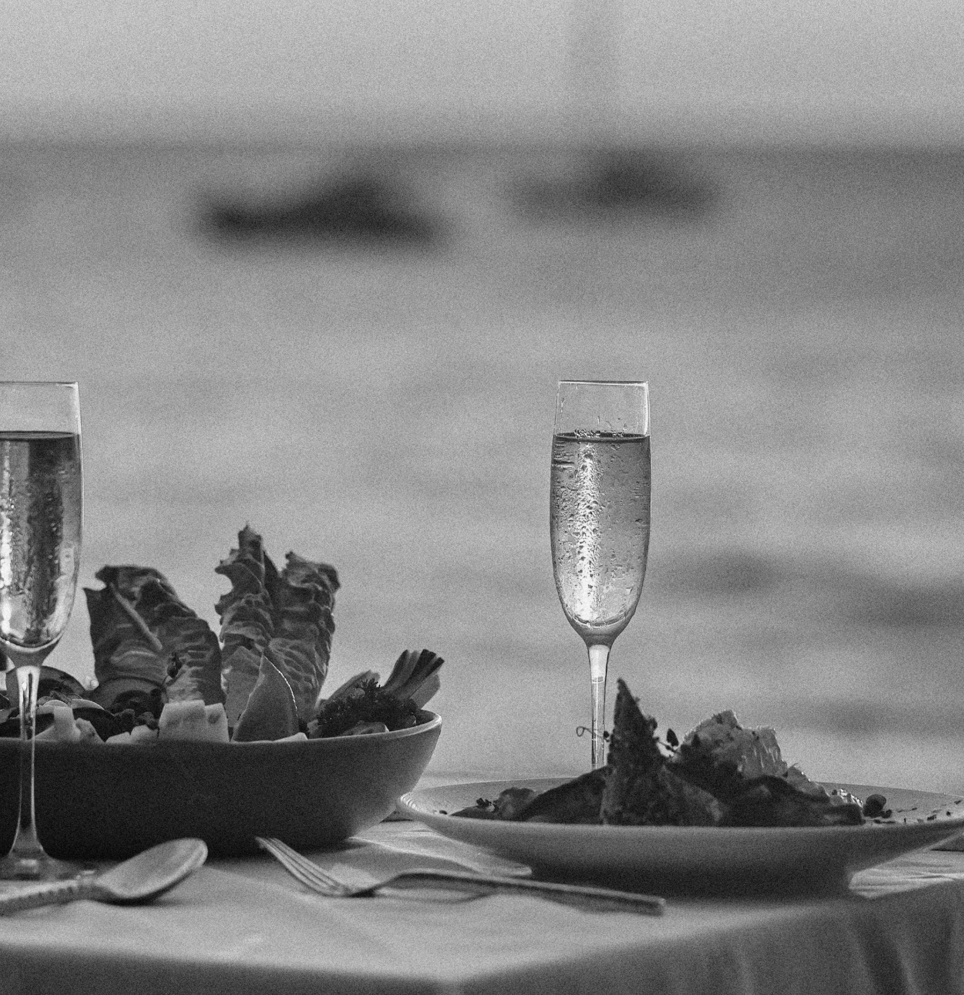 A table with potted leafy plants, a glass of water, and some scattered papers or napkins, with a high glass of bubbly drink in the background, set against a plain wall.