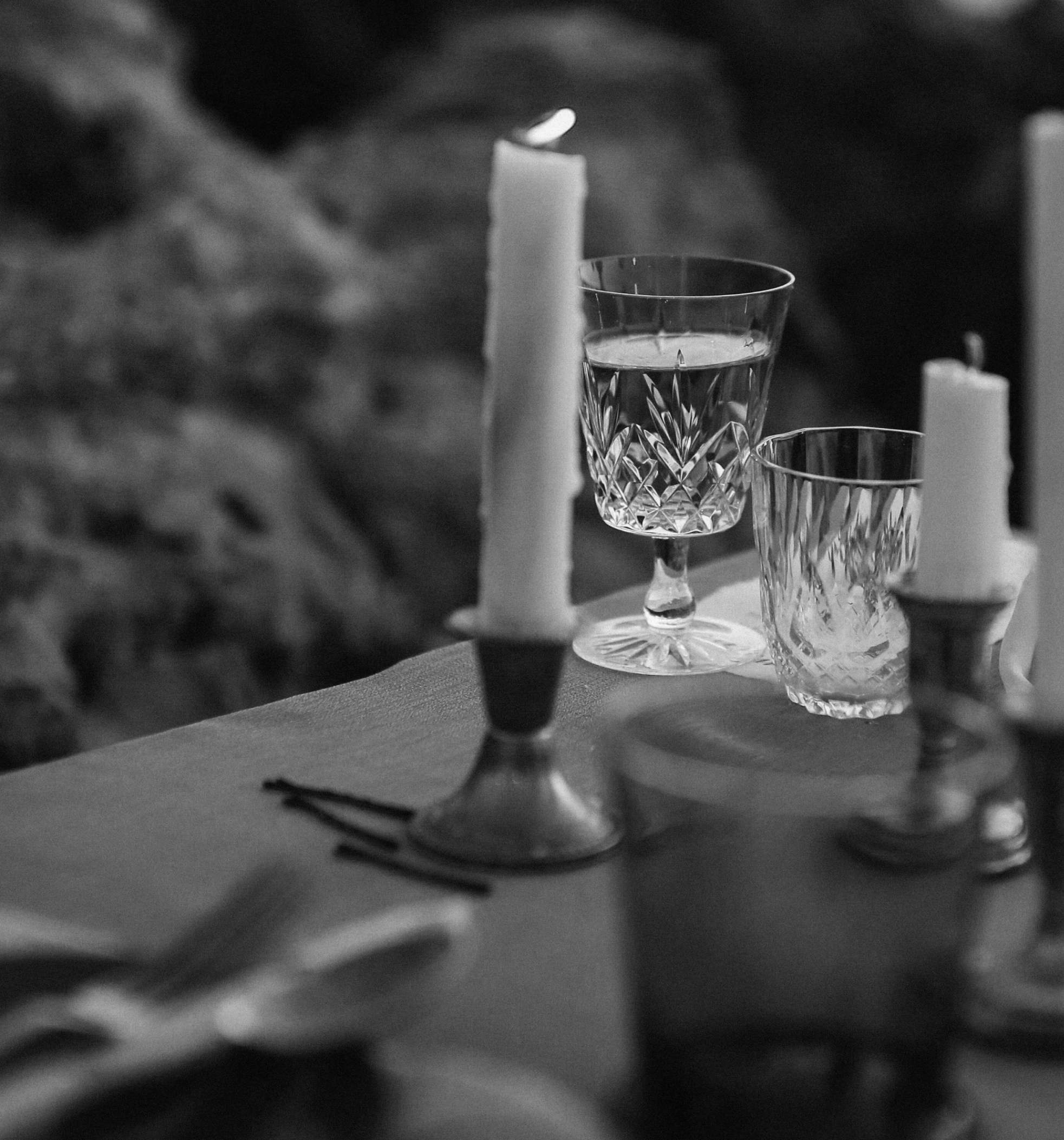 Black and white photo of a glass filled with water, placed on a surface near a candle and other glasses.