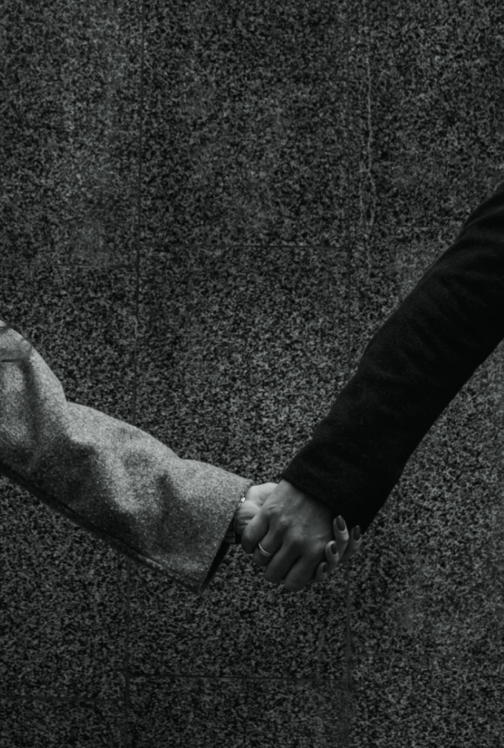 Close-up of two people holding hands against a textured stone wall surface, one person in a gray coat and the other in a black coat.