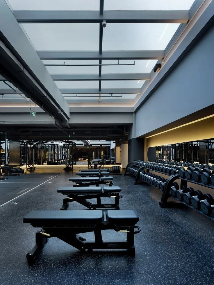 Empty gym with workout benches, dumbbells on racks, and large windows overhead.