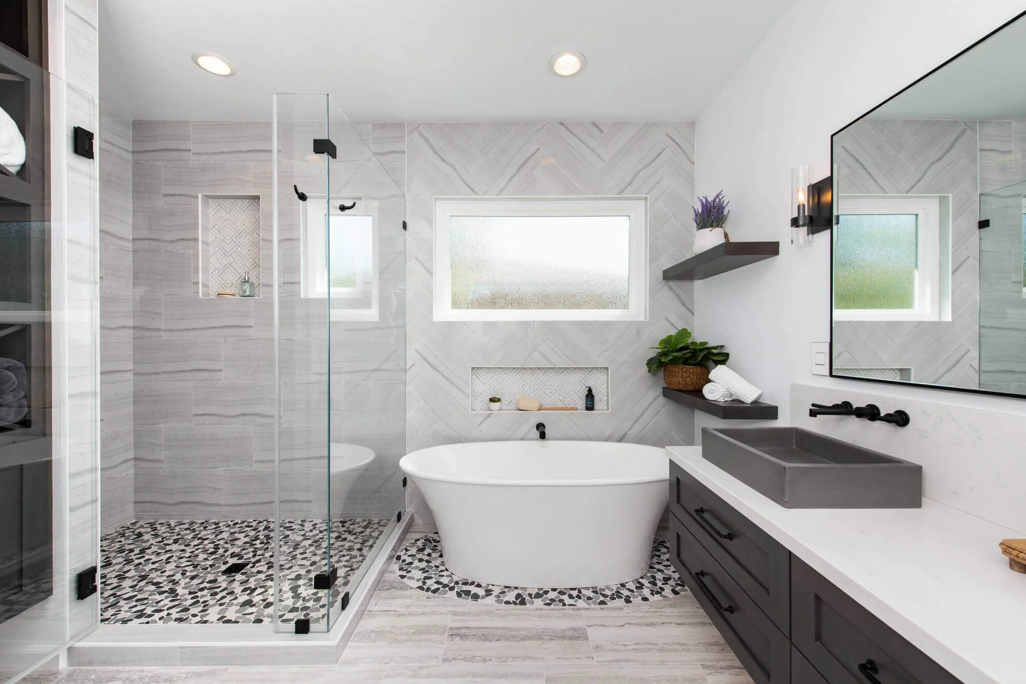 Spa-inspired bathroom remodel with freestanding soaking tub, glass walk-in shower with pebble tile flooring, and herringbone tile accent wall by Mr. H Remodel in the Nashville, Tennessee area