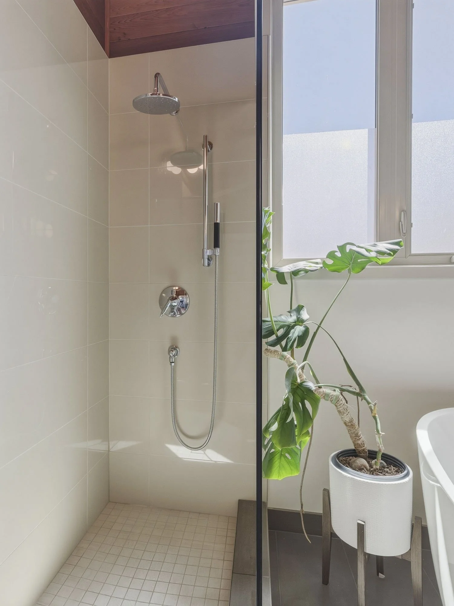 A bathroom with a walk-in shower, beige tiled walls and floor, a window with frosted glass, and a large potted plant.
