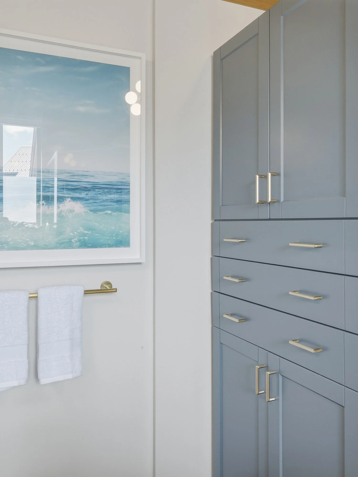 A room with a large window showing the ocean and sky outside, gray kitchen cabinets with gold handles, and two white towels hanging on a gold towel bar.