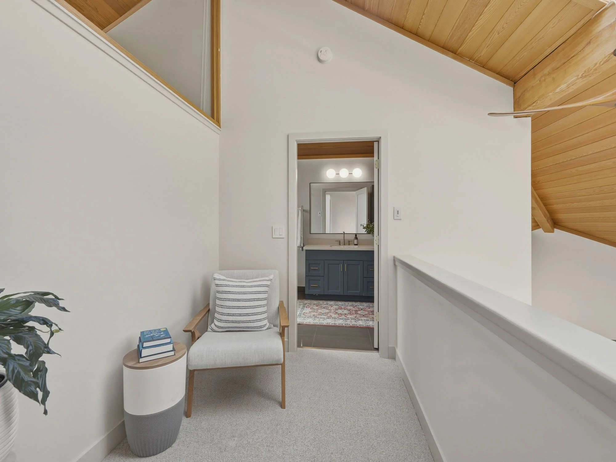 Interior of a modern home with a white wall, a wood ceiling, and a doorway leading to a bathroom with a blue vanity and mirror. There is a cozy chair with a striped pillow, a small side table with books, and a potted plant.