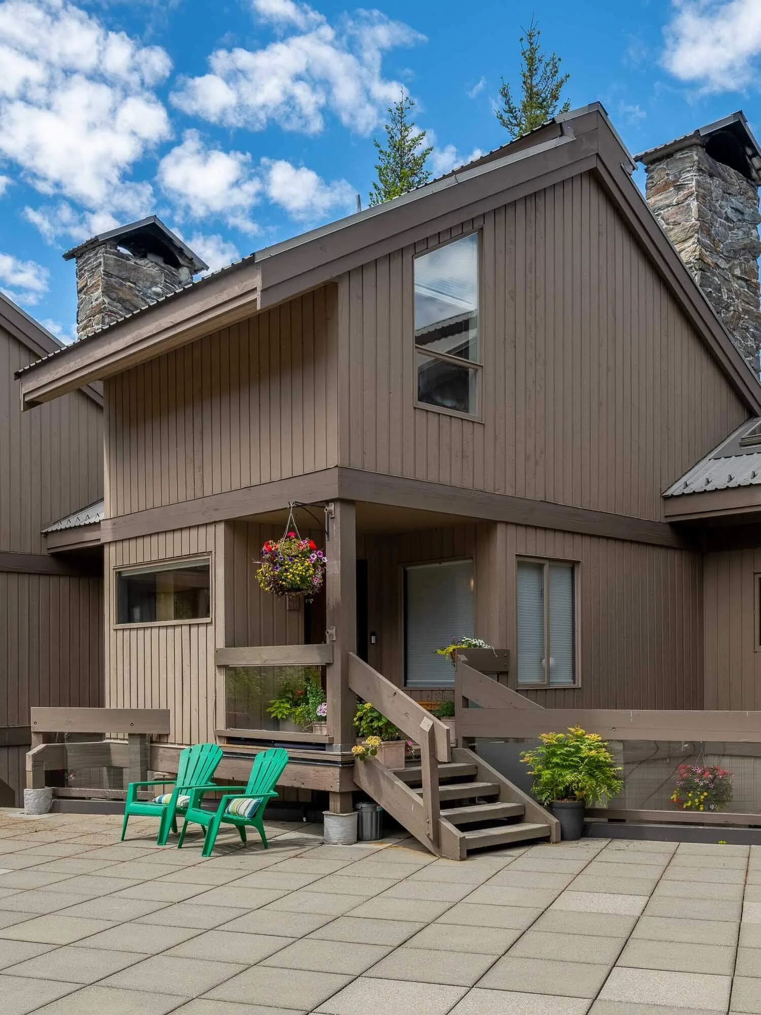 Backyard patio area with beige wooden house featuring stairs, a deck with hanging flower baskets, and two green Adirondack chairs on a tiled patio, with a stone chimney and blue sky in the background.