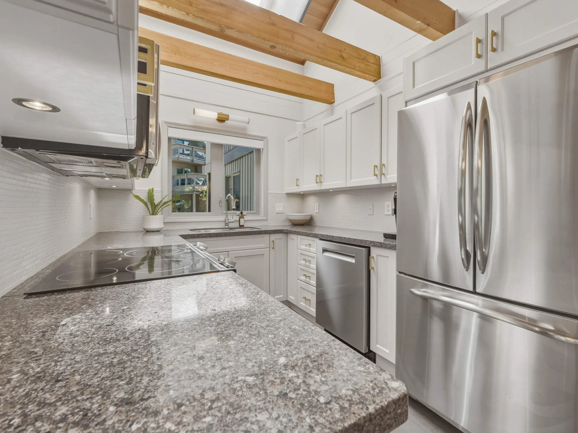 Bright kitchen at 205-6117 Eagle Drive featuring clean white cabinetry, solid stone countertops, premium stainless steel appliances, and a large window looking out toward the complex greenery.
