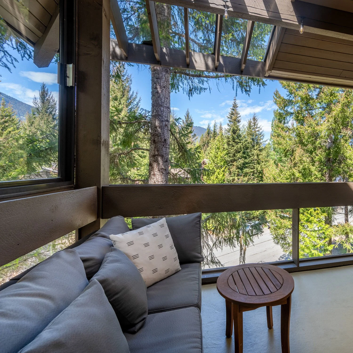 A cozy outdoor balcony or patio area with a gray couch and cushions, surrounded by large windows and overlooking green trees and mountains under a blue sky.