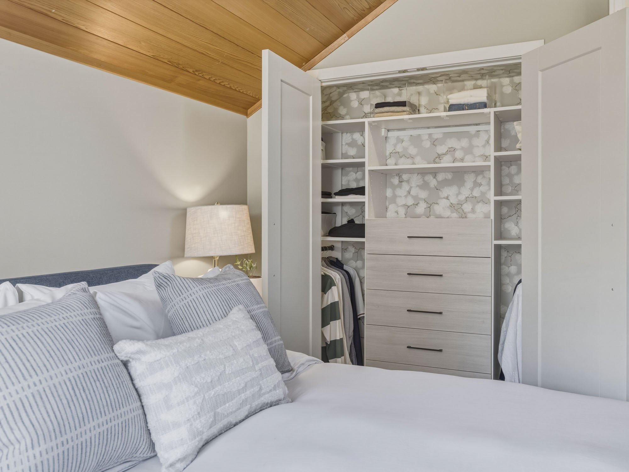 A bedroom with a bed covered in white and gray striped pillows, a white lamp, and an open closet with folded clothes and some hanging garments.