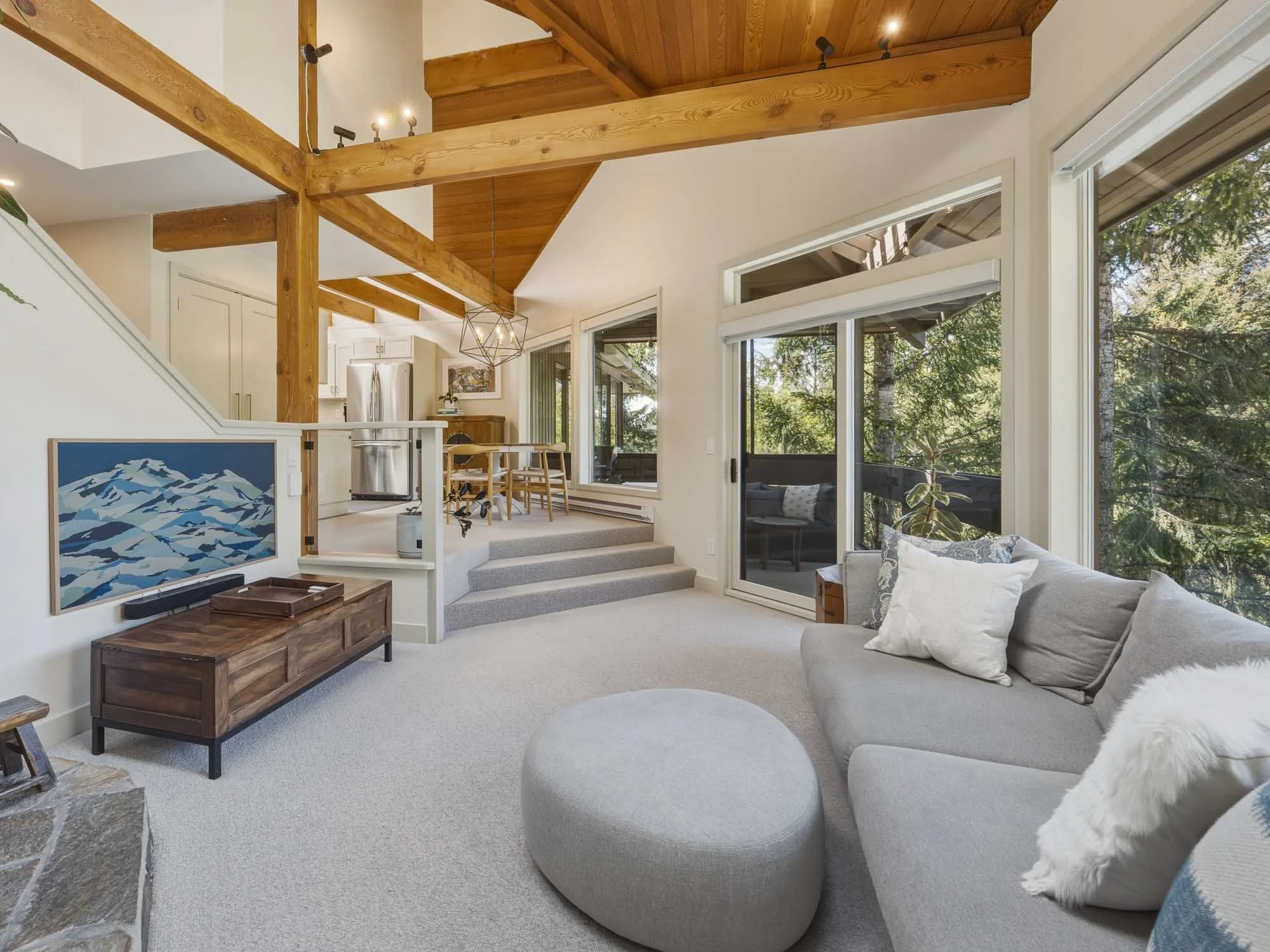 Main floor living area of this Whistler 2-bedroom townhouse for sale, highlighting expansive floor-to-ceiling windows, mountain artwork, and the open staircase leading to the private primary suite.