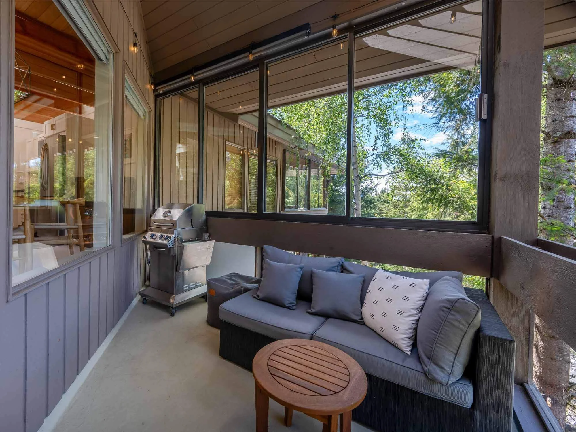 Enclosed balcony with large windows, grey couch with pillows, a small round wooden side table, a gas grill, and outdoor string lights, overlooking green trees and blue sky.